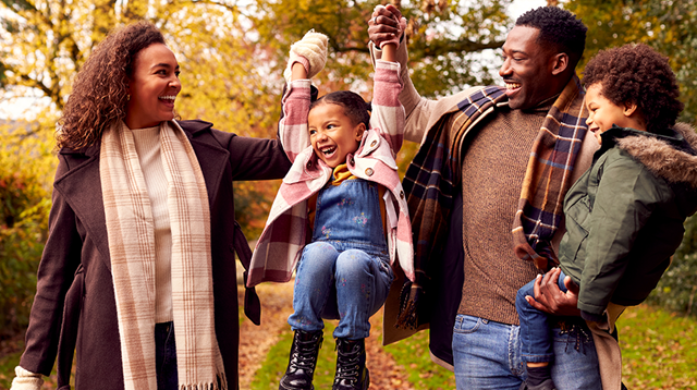 Smiling family of four with dad holding his son on his hip with dad and mum swinging the daughter between them in an autumnal walk